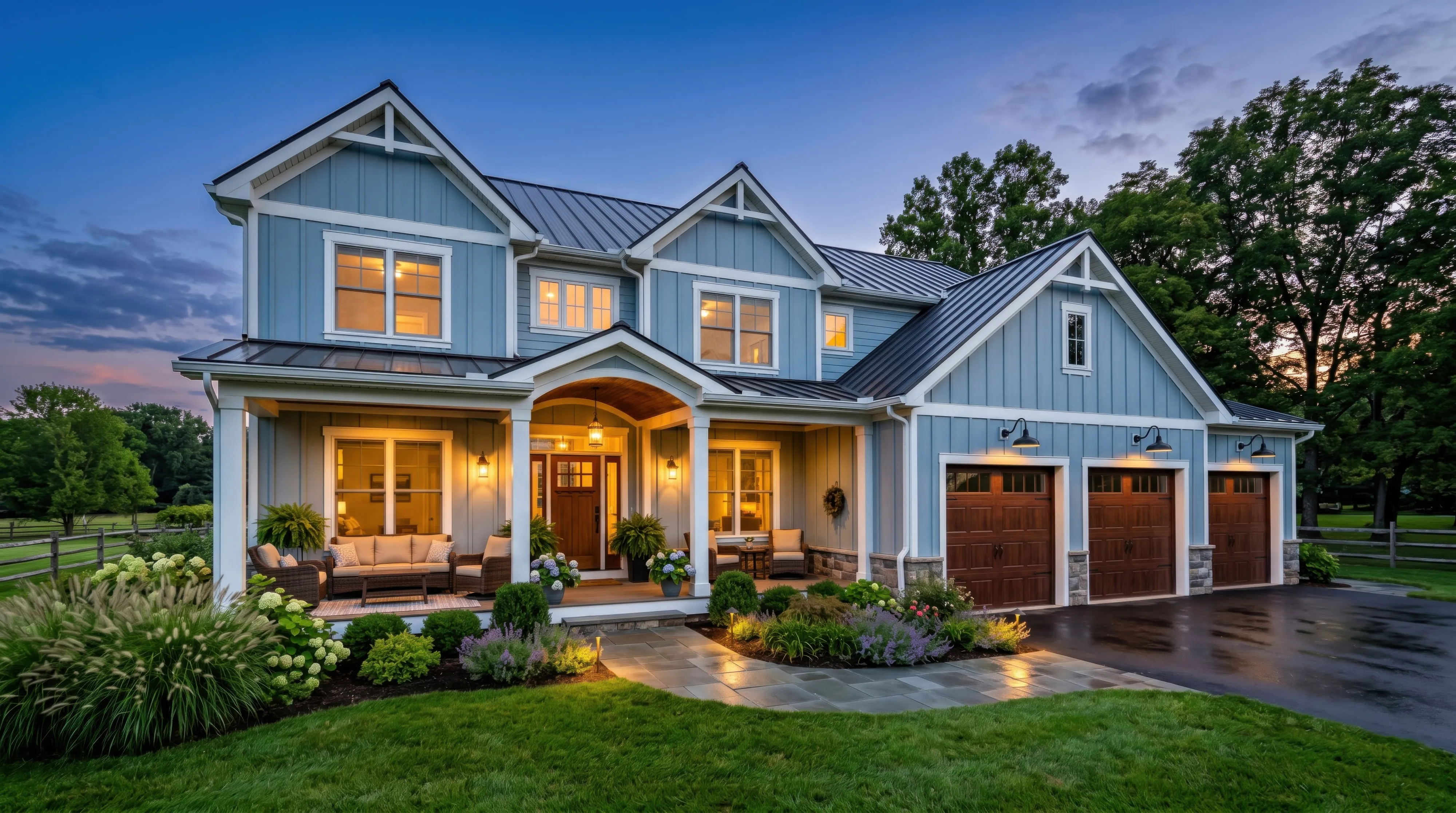 A blue board-and-batten farmhouse at dusk with a covered front porch, lantern lighting and a three-bay garage, framed by mature trees.