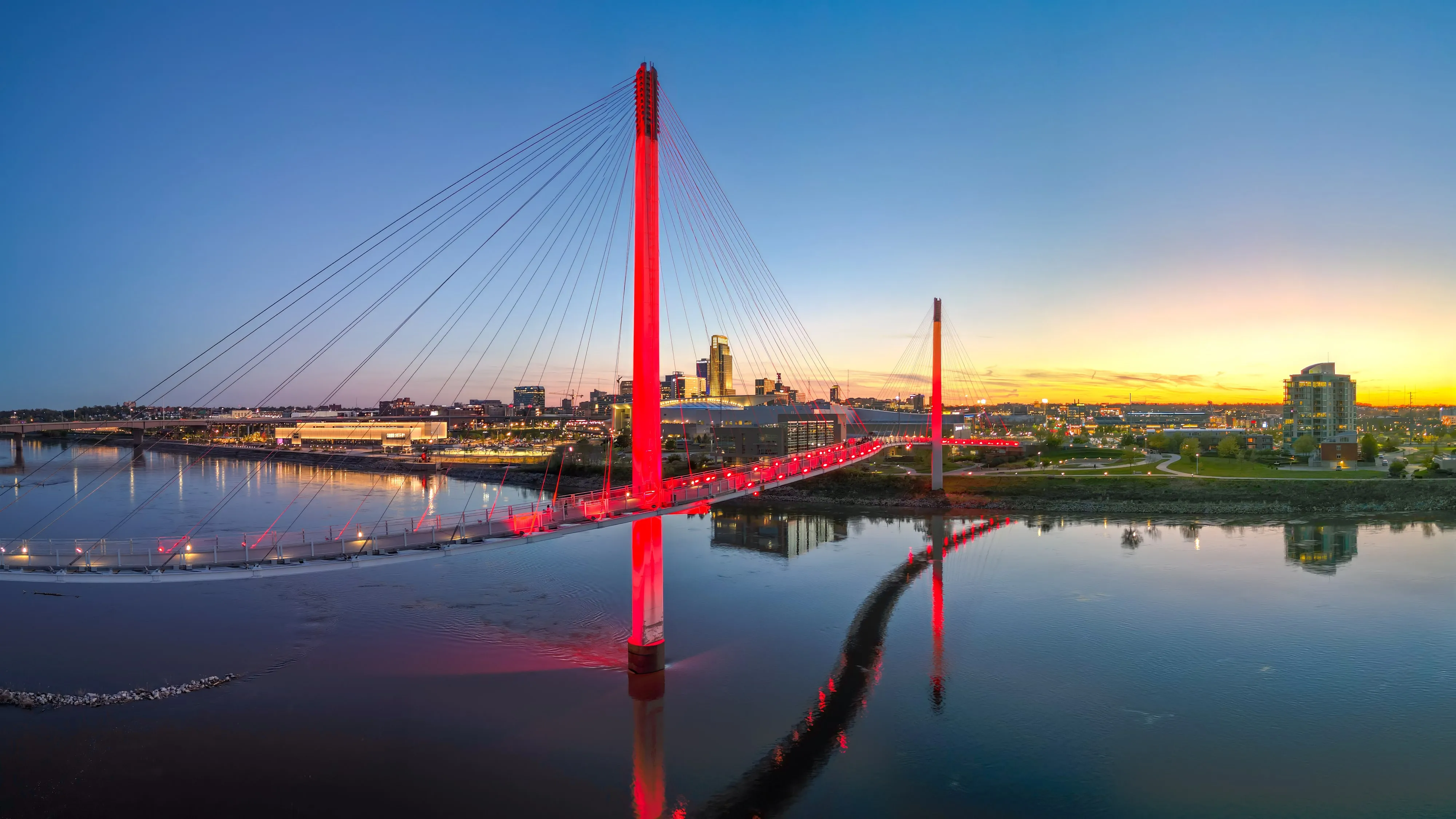 The Bob Kerrey Pedestrian Bridge lit red at sunset, its cables reflected in the calm Missouri River with the downtown Omaha skyline beyond.