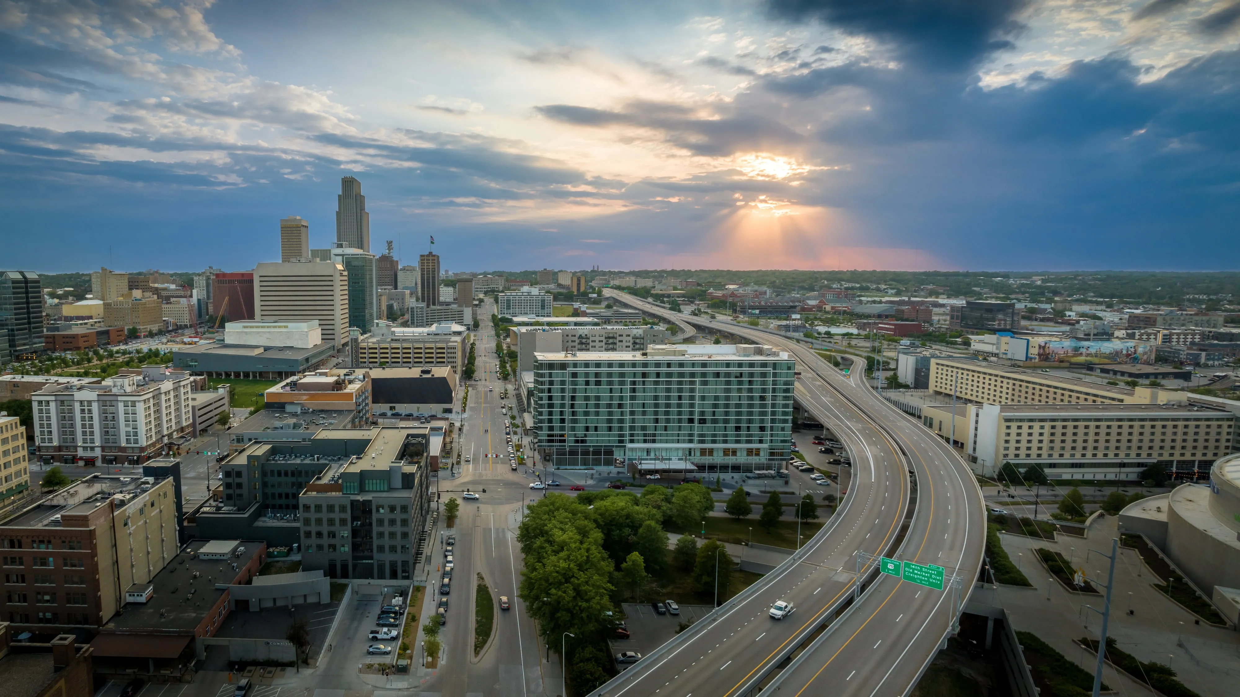 An aerial view of downtown Omaha at sunset, looking along Dodge Street past the high-rises as sunlight breaks through the clouds.