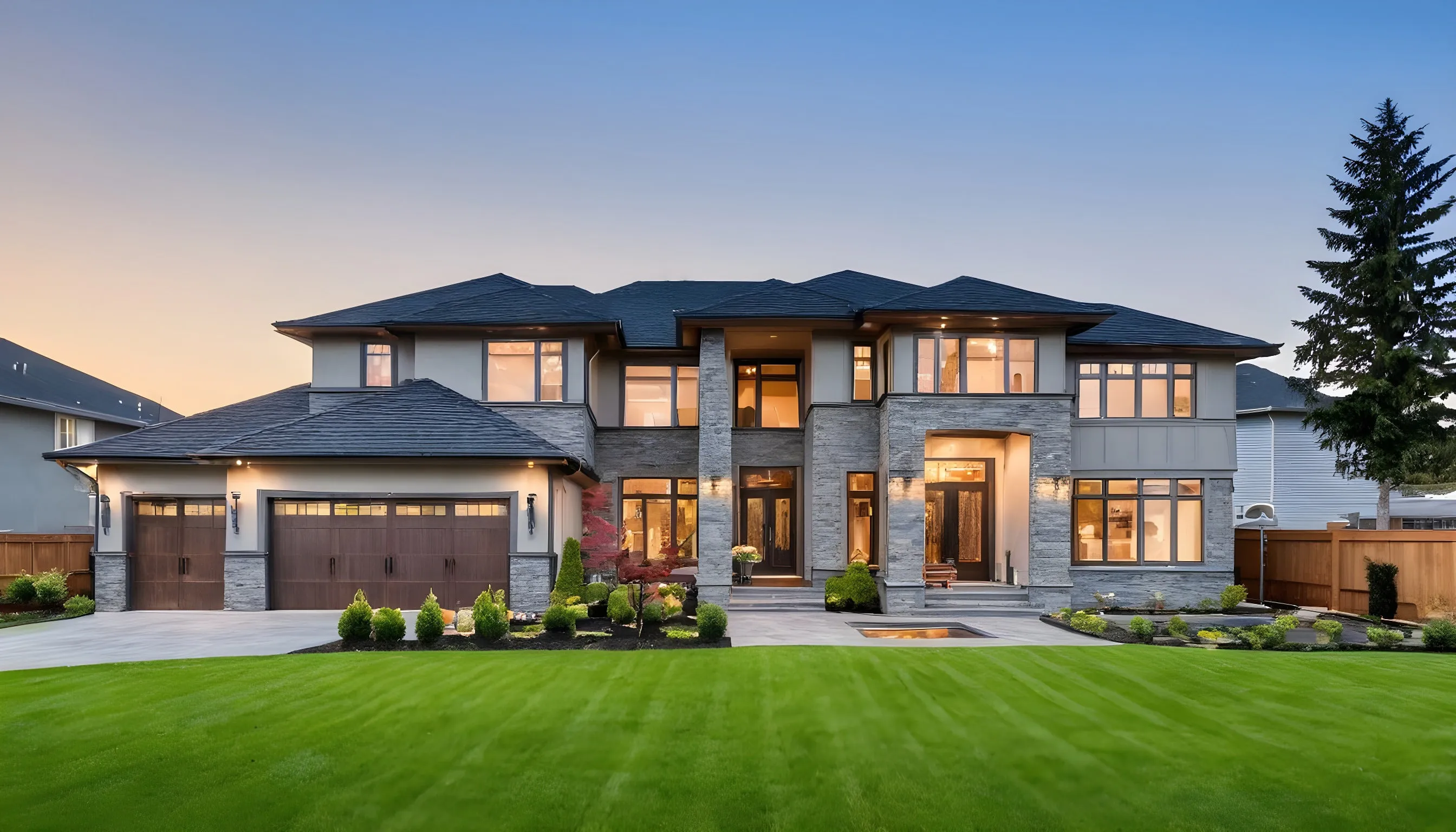 A two-story stone and stucco home at twilight, warmly lit from within, set behind a deep manicured lawn with a three-car garage.