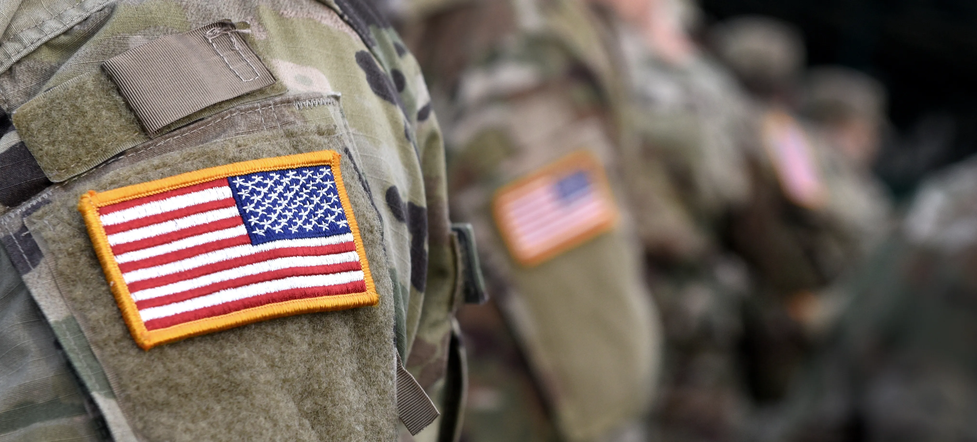 A United States flag patch on the sleeve of a camouflage uniform, with a line of service members out of focus behind.